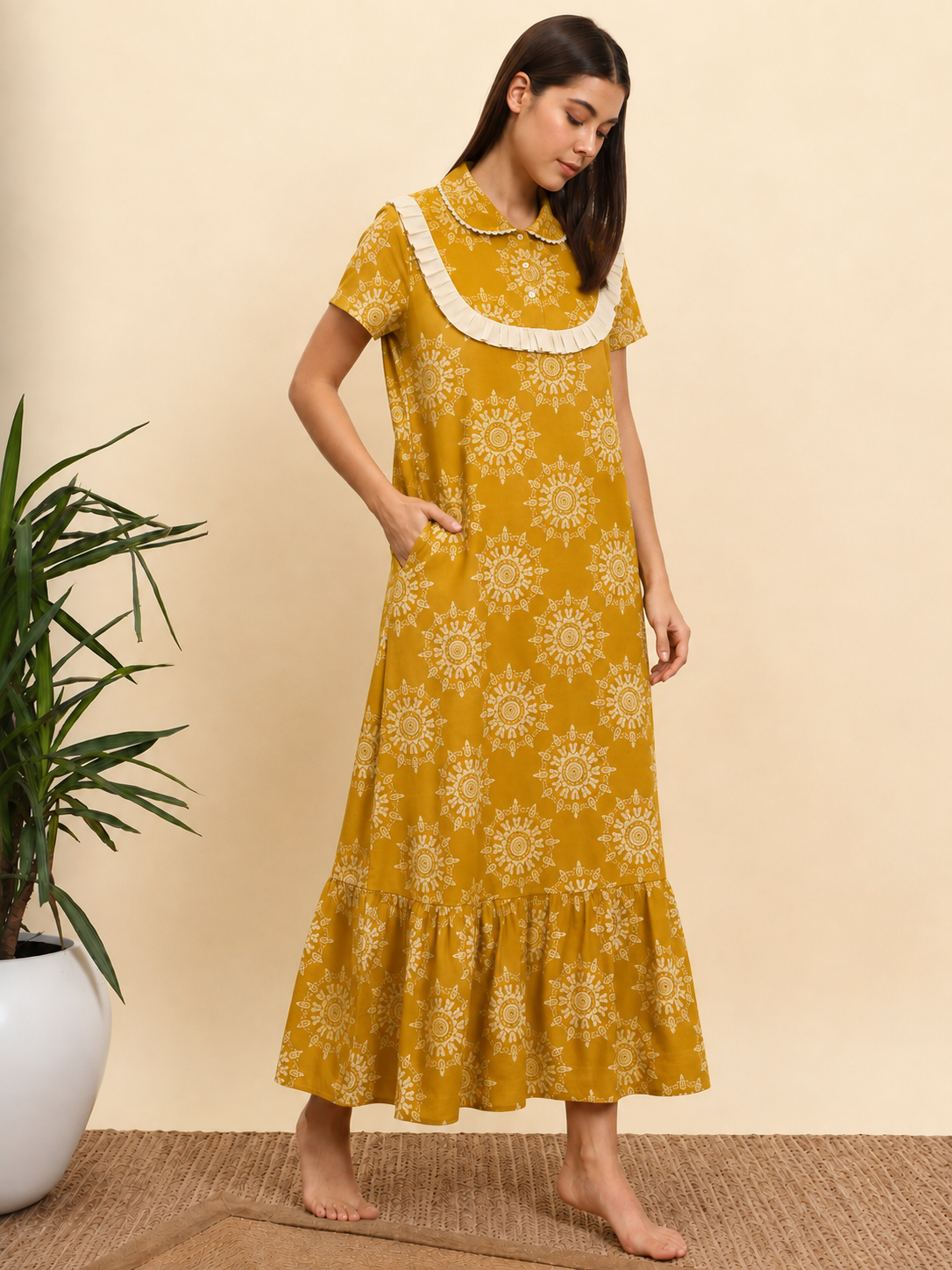 A woman wearing a mustard yellow midi dress with a white sunburst pattern and a white lace collar, standing barefoot in a room with a beige wall and a potted plant.