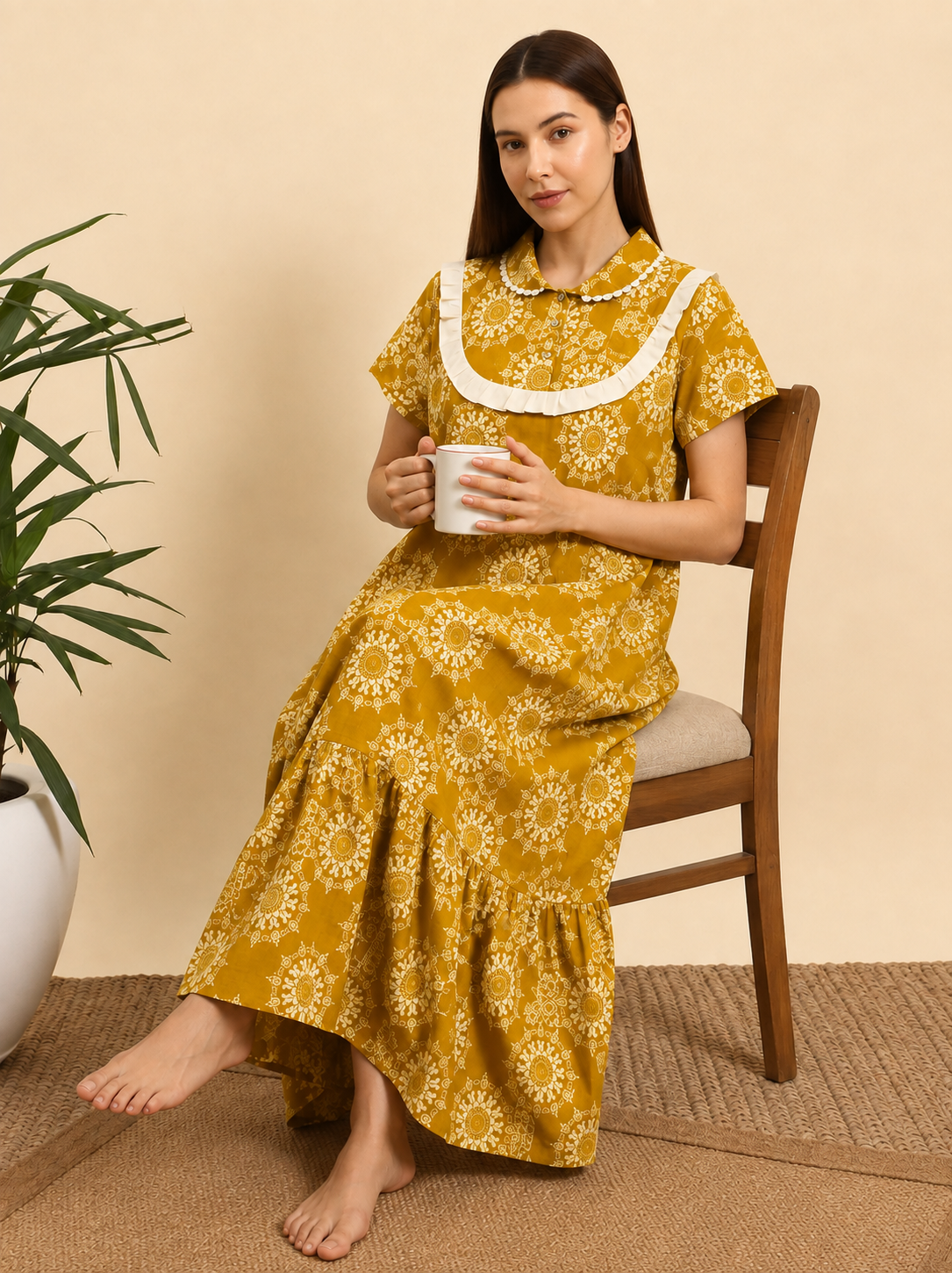 A woman sits barefoot on a wooden chair wearing a yellow floral print nightgown with a white collar, holding a white mug in a cozy indoor setting.