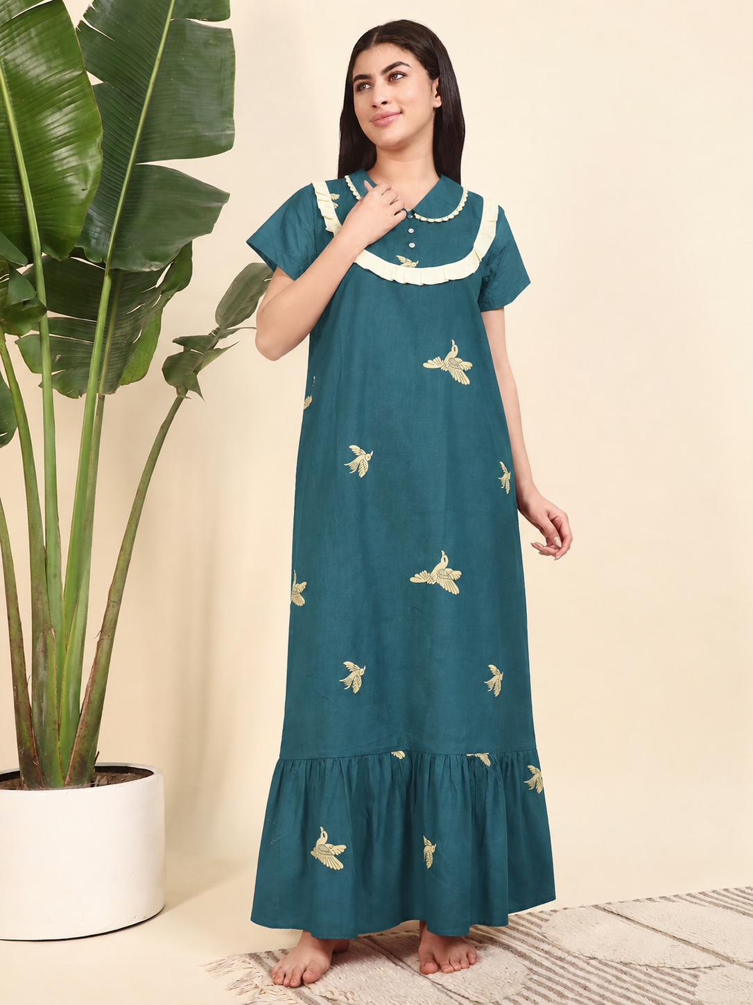 A woman modeling a teal blue maxi nightgown with white ruffled trim and small gold bird prints, standing next to a large potted plant against a beige background.