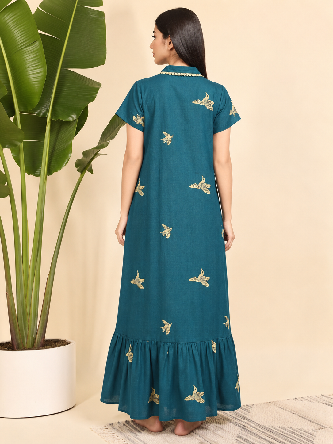 A woman stands with her back to the camera wearing a teal blue maxi dress with gold embroidered motifs. The dress features short sleeves, a ruffled hem, and a gold beaded collar.