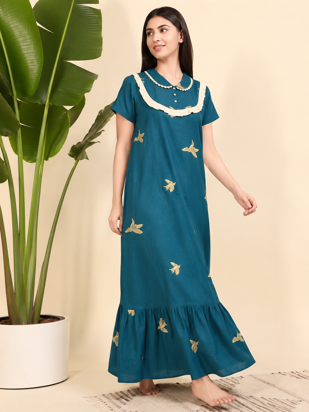 A woman wearing a teal blue maxi nightgown with gold butterfly embroidery and a cream ruffled collar, standing in a studio setting next to a potted plant.