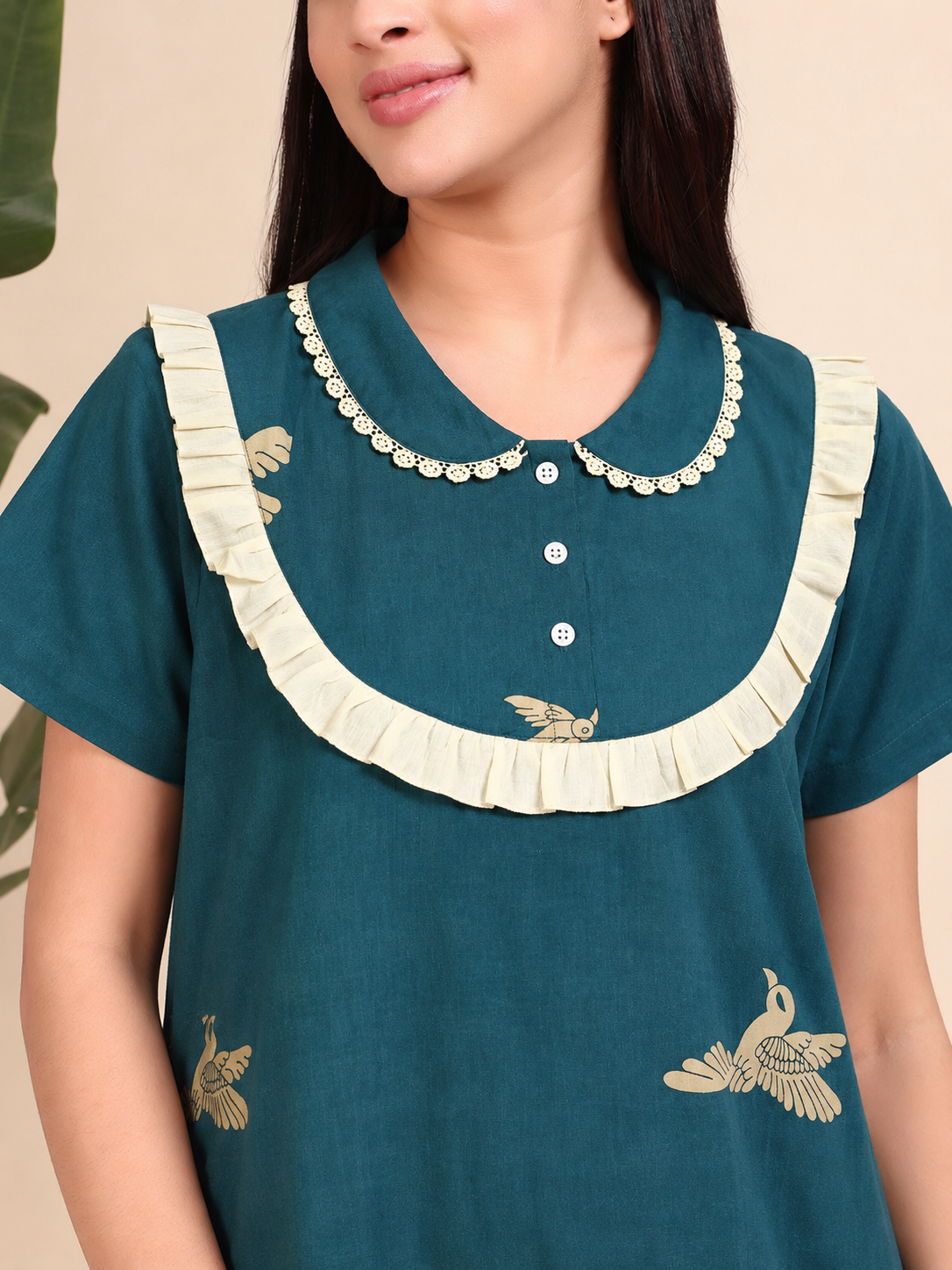 A woman wearing a teal short-sleeve tunic top with cream ruffle trim around the collar and hem, plus small beige bird embroidery on the front.