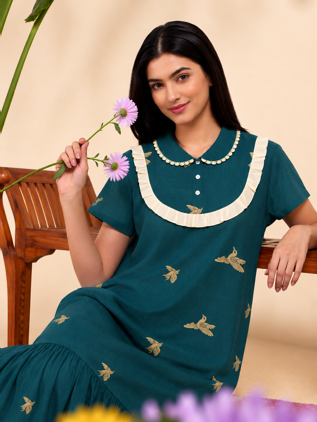 A woman wearing a teal green short-sleeve kurta dress with gold bird embroidery and a cream ruffled collar, holding a purple flower while sitting on a wooden bench.
