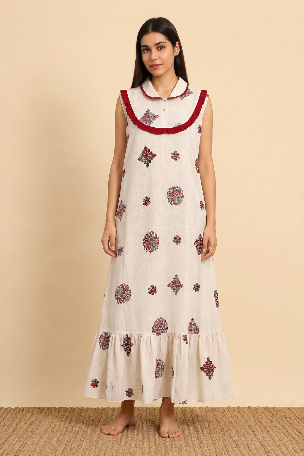 A woman stands barefoot against a warm beige studio backdrop wearing a sleeveless cream maxi dress with red embroidered border detailing and scattered red and blue floral motifs.