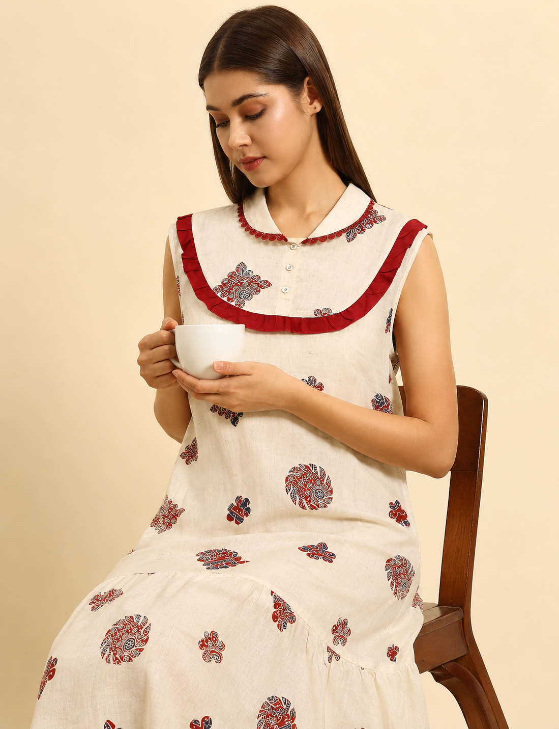 A woman seated on a wooden chair wearing a sleeveless cream-colored dress with red ruffled trim and red floral block prints, holding a white cup against a warm beige background.