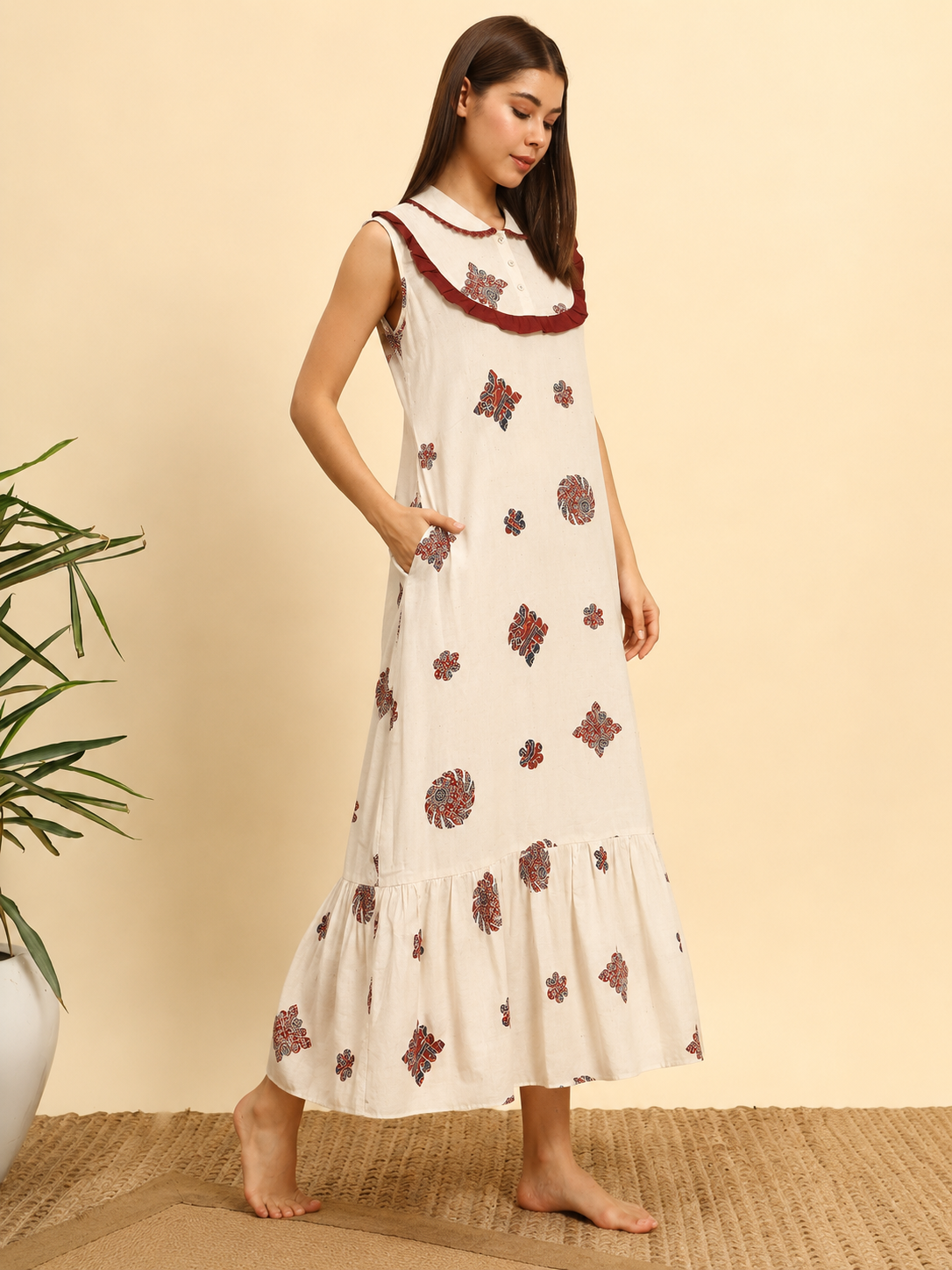 A woman modeling a sleeveless, tiered maxi dress in off-white with red and dark blue floral prints and a red ruffled collar, standing against a beige background.