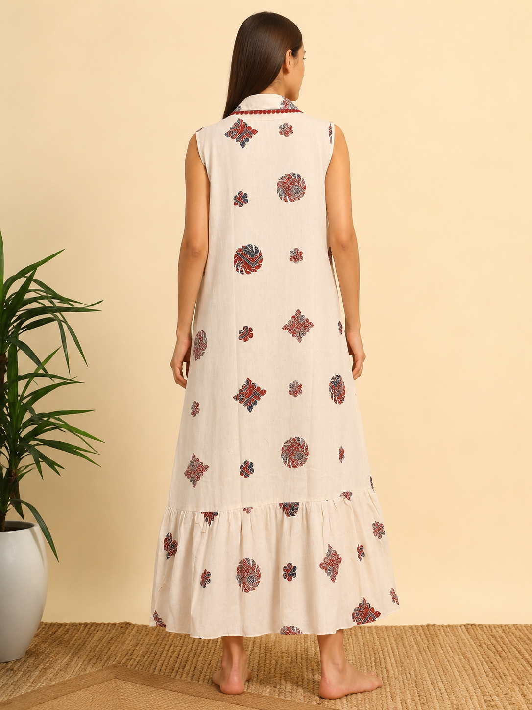 A woman stands barefoot facing away from the camera, wearing a sleeveless, tiered cream-colored dress with red and blue floral prints. The background is a solid beige wall with a potted plant on the left.