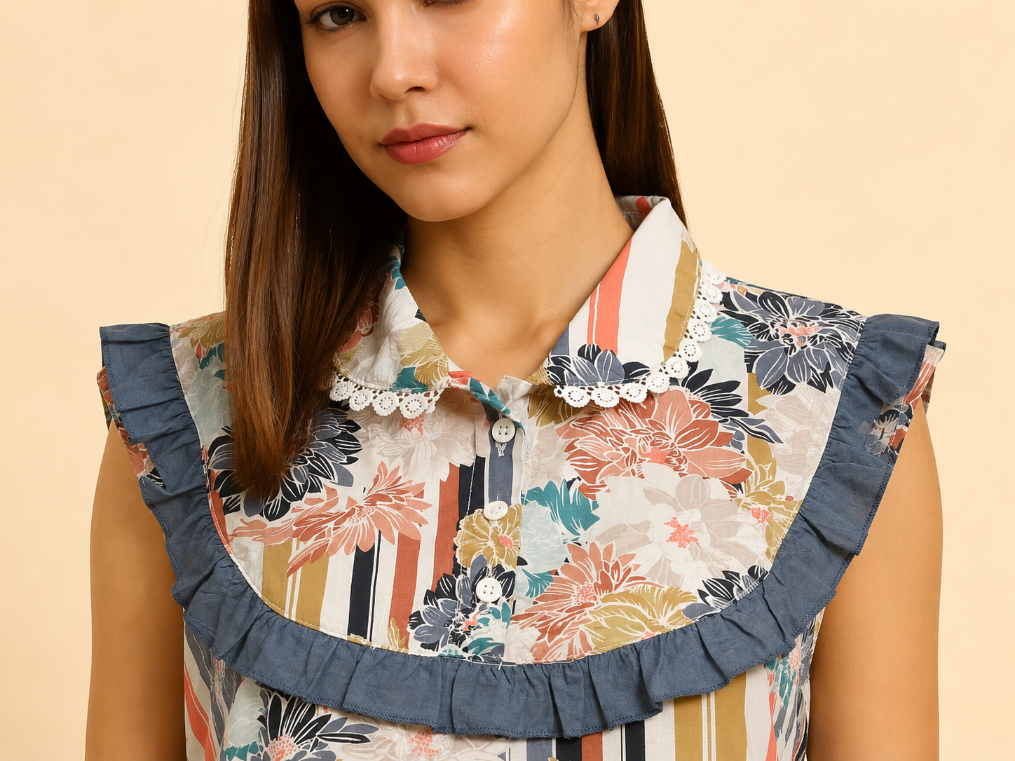 A close-up of a woman wearing a sleeveless blouse with a large ruffled collar. The blouse features a multicolor floral and striped pattern on a cream base, with a blue-gray ruffle trim and white lace trim around the collar.