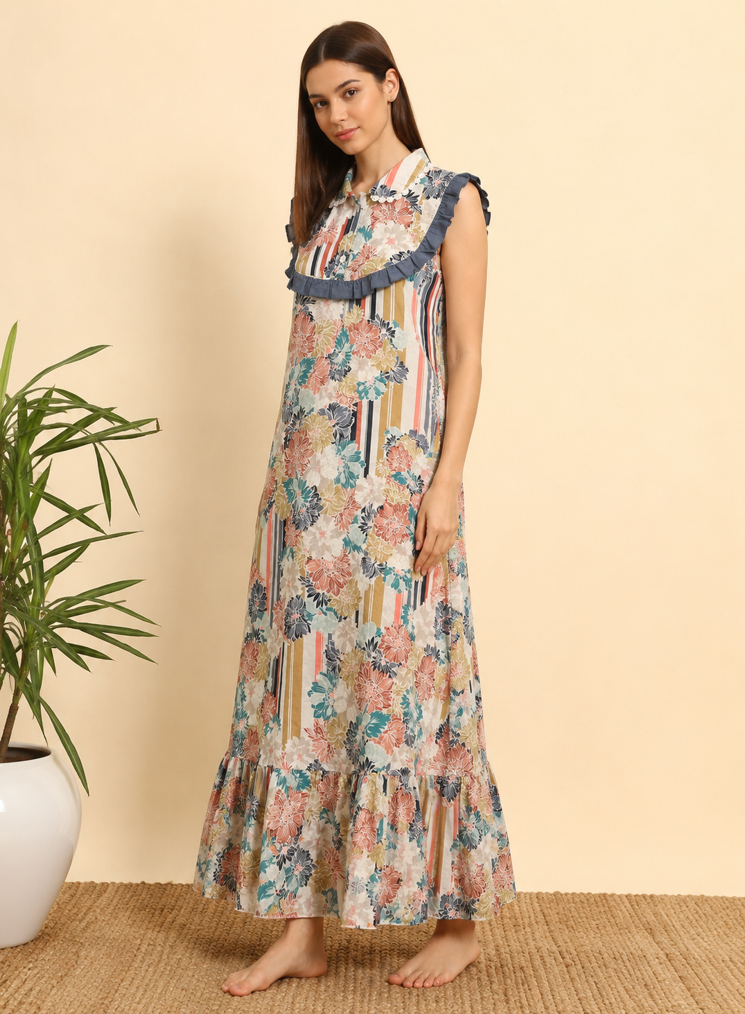A woman modeling a sleeveless, high-neck maxi dress with a multicolor floral and striped pattern, ruffled trim at the neckline, and ruffled hem, standing barefoot on a woven rug next to a potted plant.