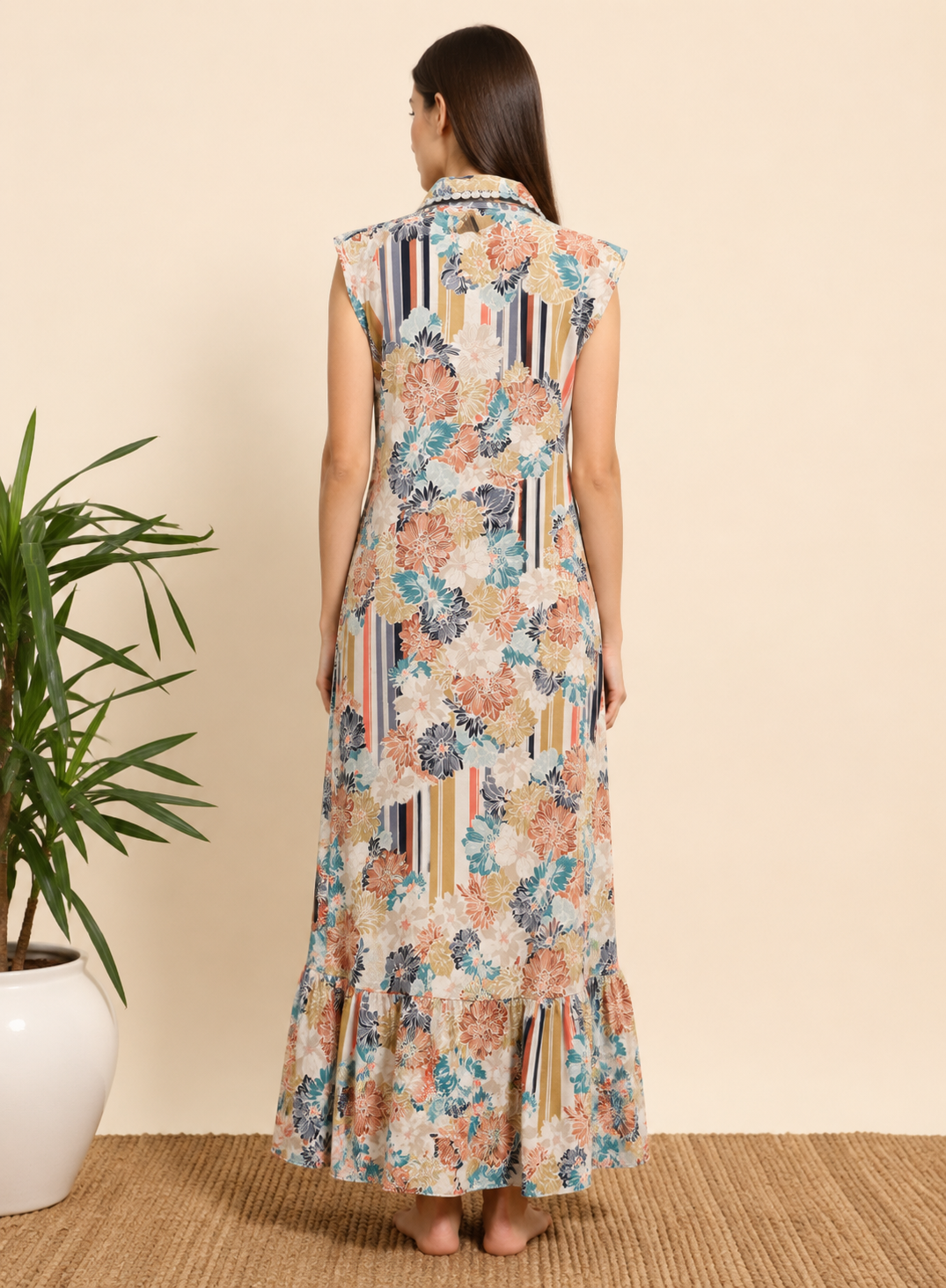 A woman viewed from behind wearing a sleeveless maxi dress with a multicolor floral and striped pattern, standing on a woven rug against a beige wall next to a potted plant.