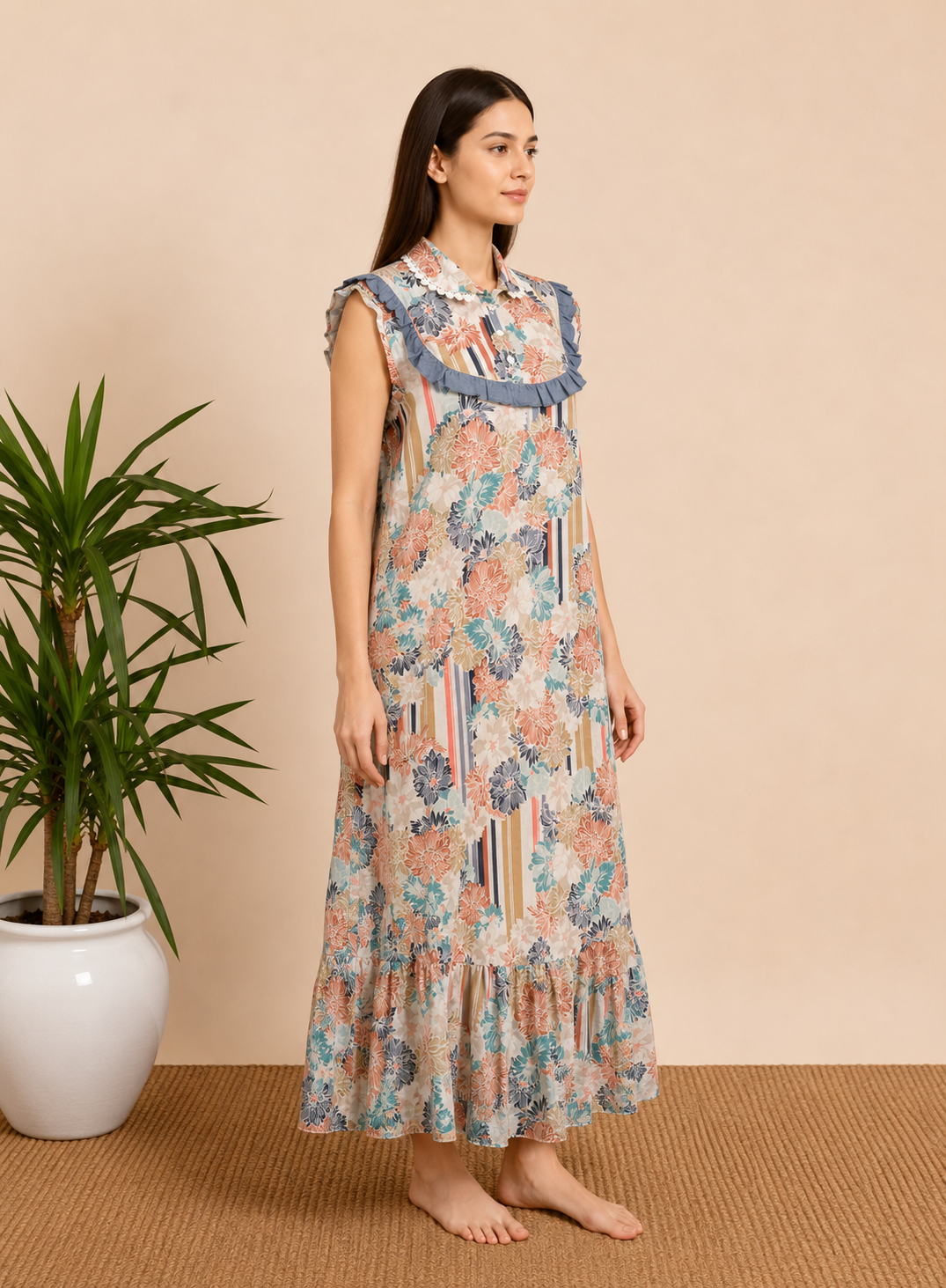 A woman stands barefoot in a studio setting wearing a sleeveless, ruffled maxi dress with a multicolor floral and striped print. A potted plant is visible on the left against a warm beige backdrop.
