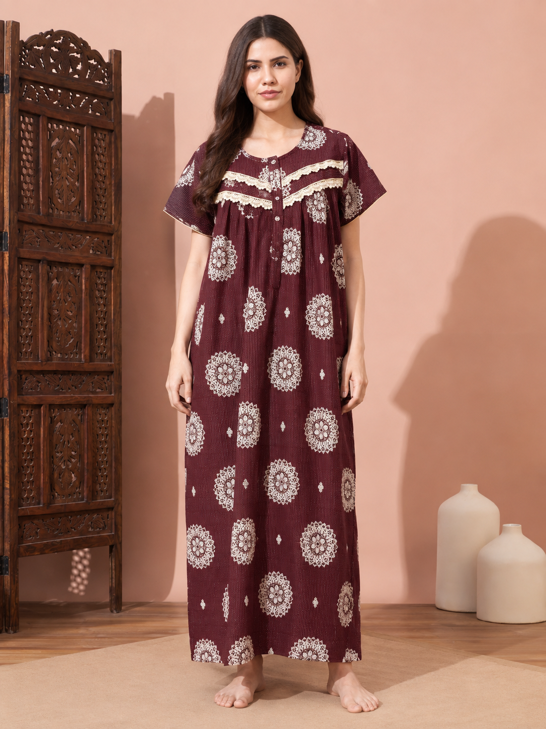 A woman stands barefoot in a studio setting wearing a long maroon nightgown with white circular mandala prints and cream lace trim at the neckline. A carved wooden screen is on the left, and two white ceramic vases sit on the floor against a peach-colored wall.