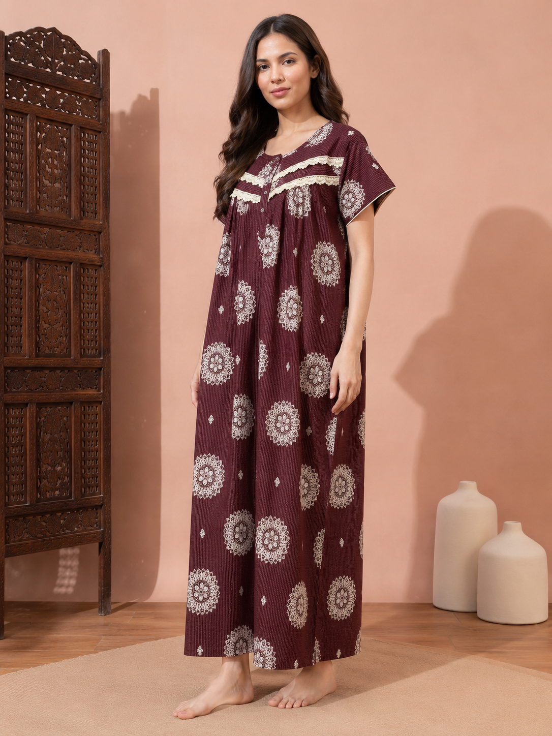 A woman stands barefoot in a room wearing a long maroon nightgown with cream circular floral prints and cream lace trim at the neckline. A carved wooden cabinet is on the left, and two white ceramic vases are on the floor against a peach-colored wall.