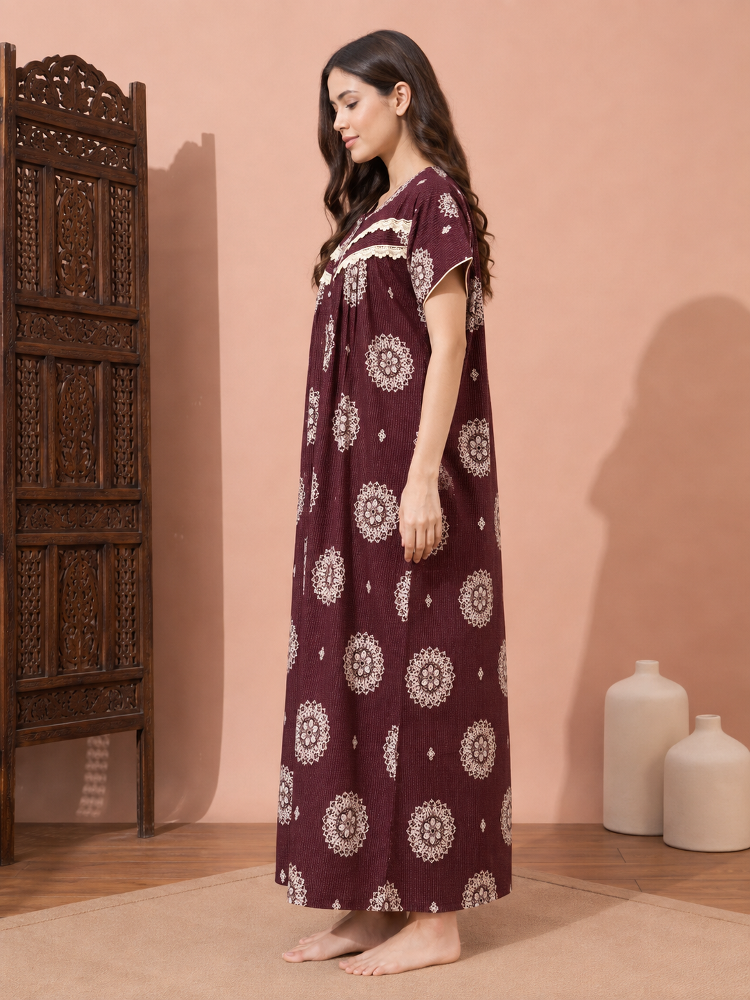 A woman stands barefoot in profile wearing a long, loose-fitting maroon nightgown with cream circular floral prints and a cream embroidered neckline. The setting features a warm peach-pink wall, a carved dark wood screen on the left, and two white ceramic vases on the right.