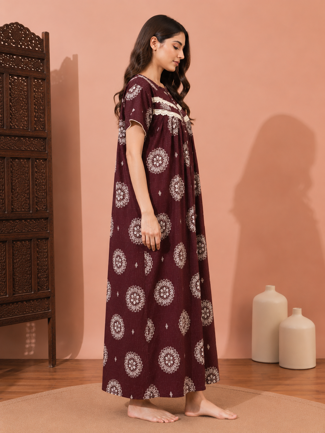 A woman stands barefoot in a studio setting wearing a long maroon nightgown with white circular mandala patterns and white embroidered detailing at the neckline. The background is a solid peach-pink wall with a dark wooden screen on the left and two white ceramic vases on the right.