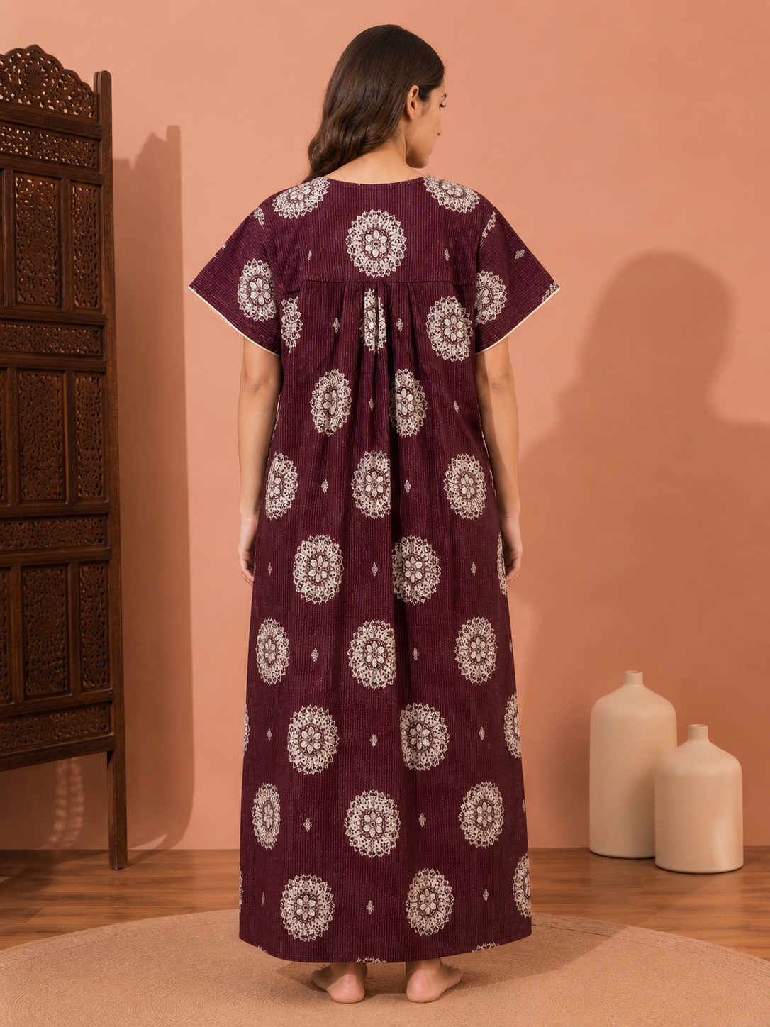 A woman stands with her back to the camera wearing a long, loose-fitting maroon nightgown or tunic dress with white circular mandala patterns. The setting is a warm-toned room with a peach-pink wall, a dark carved wooden cabinet on the left, and two white ceramic vases on the floor to the right.
