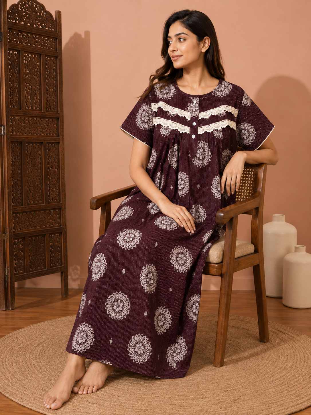 A woman sits on a wooden chair wearing a long, short-sleeved nightgown with a deep purple base and white floral mandala patterns. The setting features a warm peach-colored wall, a carved wooden screen, and neutral-toned ceramic vases.