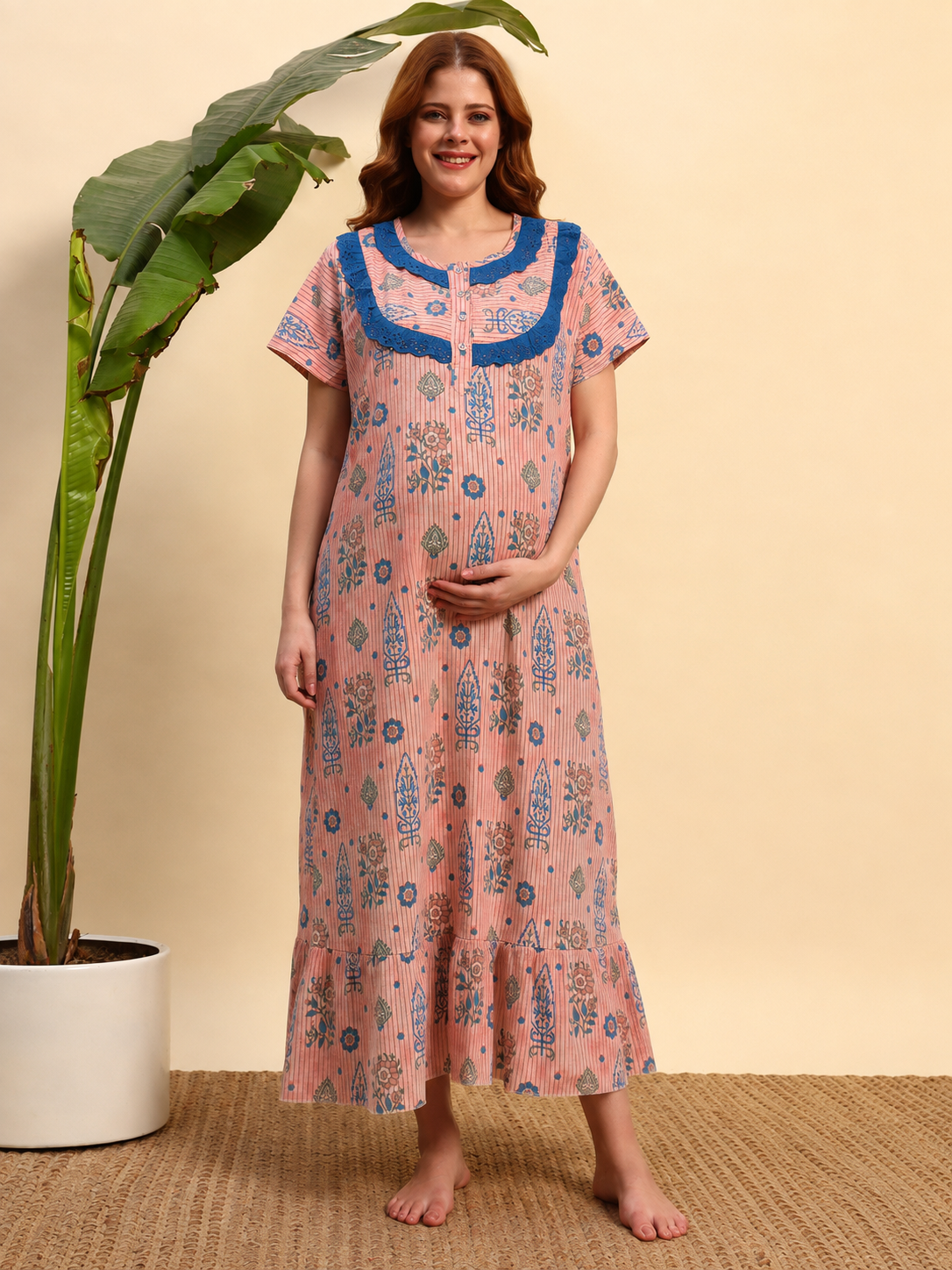 A woman stands barefoot indoors wearing a pink printed nightgown with blue trim and blue floral motifs. She is posed next to a large potted plant against a warm beige wall.