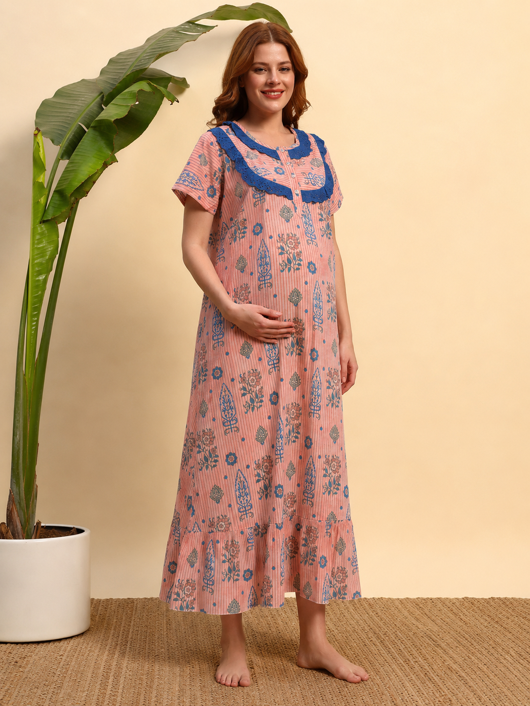 A woman wearing a pink long nightgown with blue floral prints and a blue collar, standing barefoot in front of a beige wall next to a potted plant.