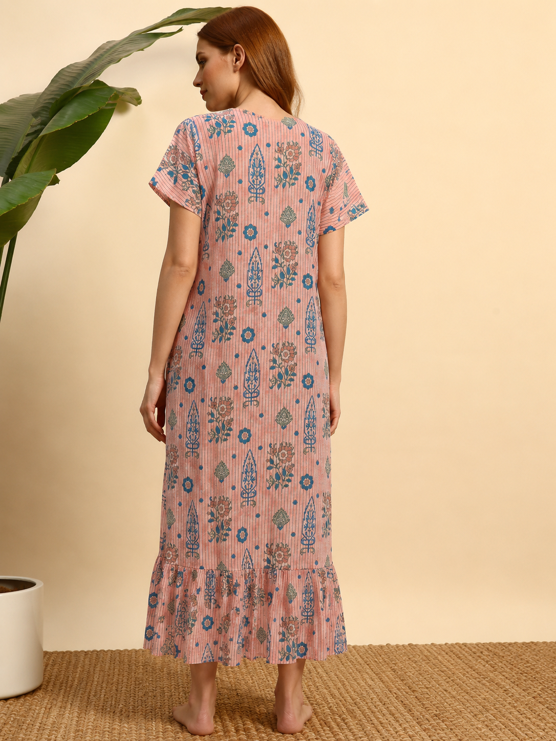 A woman stands barefoot indoors wearing a pink short-sleeve maxi dress with blue and beige floral and geometric patterns, photographed from the back against a warm beige wall with a plant and woven rug visible.