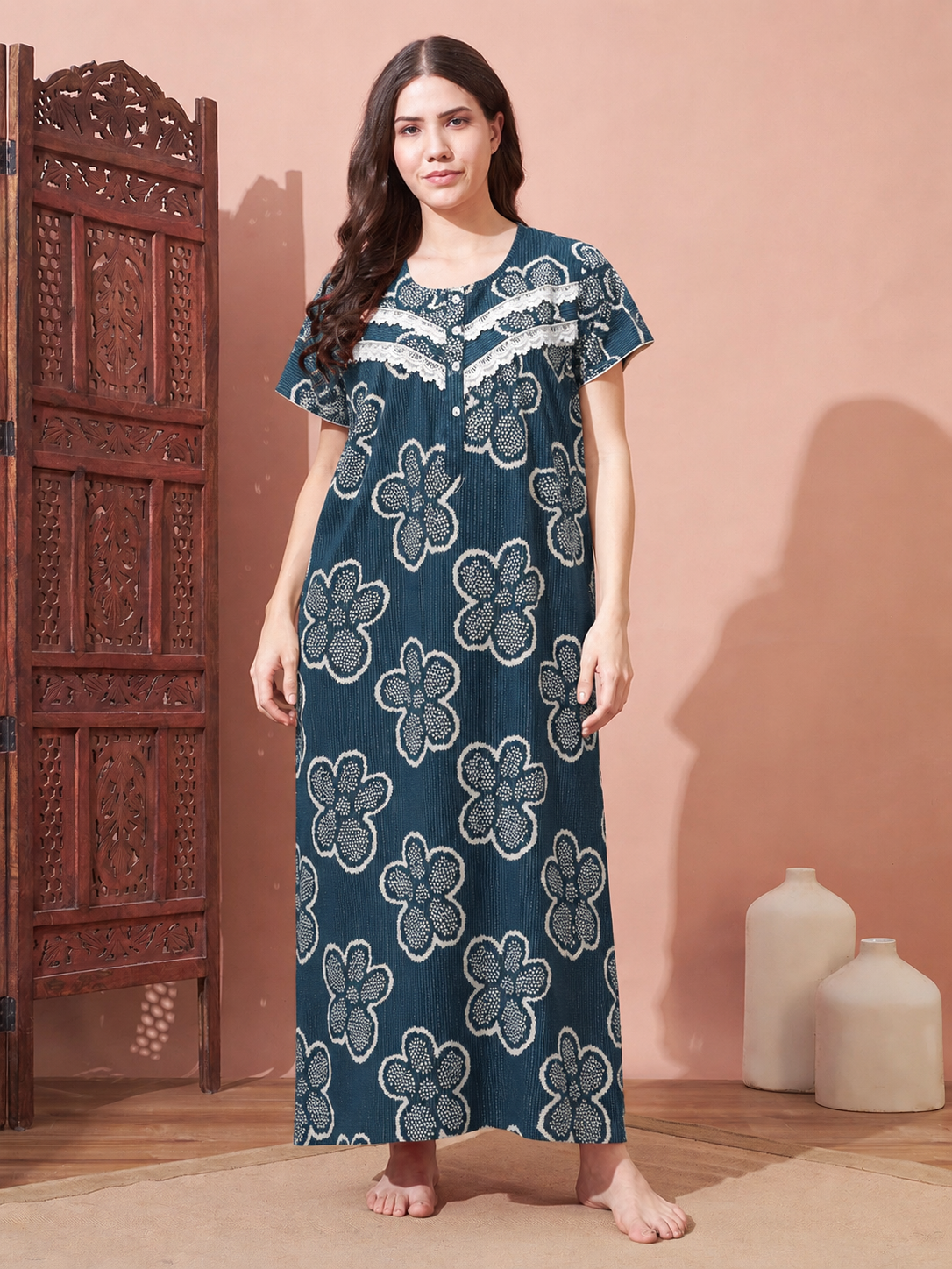 A woman stands barefoot in a room wearing a long teal nightgown with a white floral block print pattern and lace trim at the neckline. She is posed next to a carved wooden screen and ceramic vases against a warm peach-colored wall.