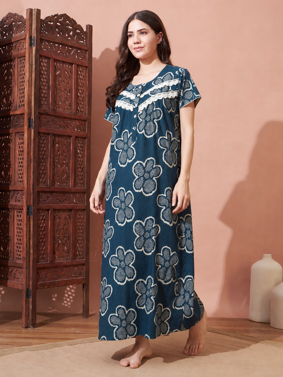 A woman stands barefoot in a room wearing a long teal nightgown with a large floral block-print pattern in cream and blue-gray. The background features a peach-colored wall, a carved wooden folding screen, and a light-colored ceramic vase.