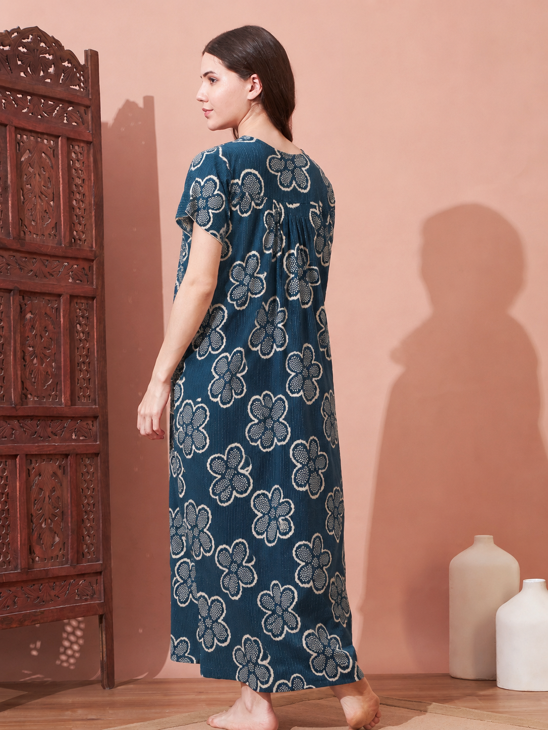 A woman stands barefoot indoors wearing a long-sleeve maxi dress with a blue and white floral block-print pattern, photographed against a peach-pink wall with a carved wooden screen on the left.