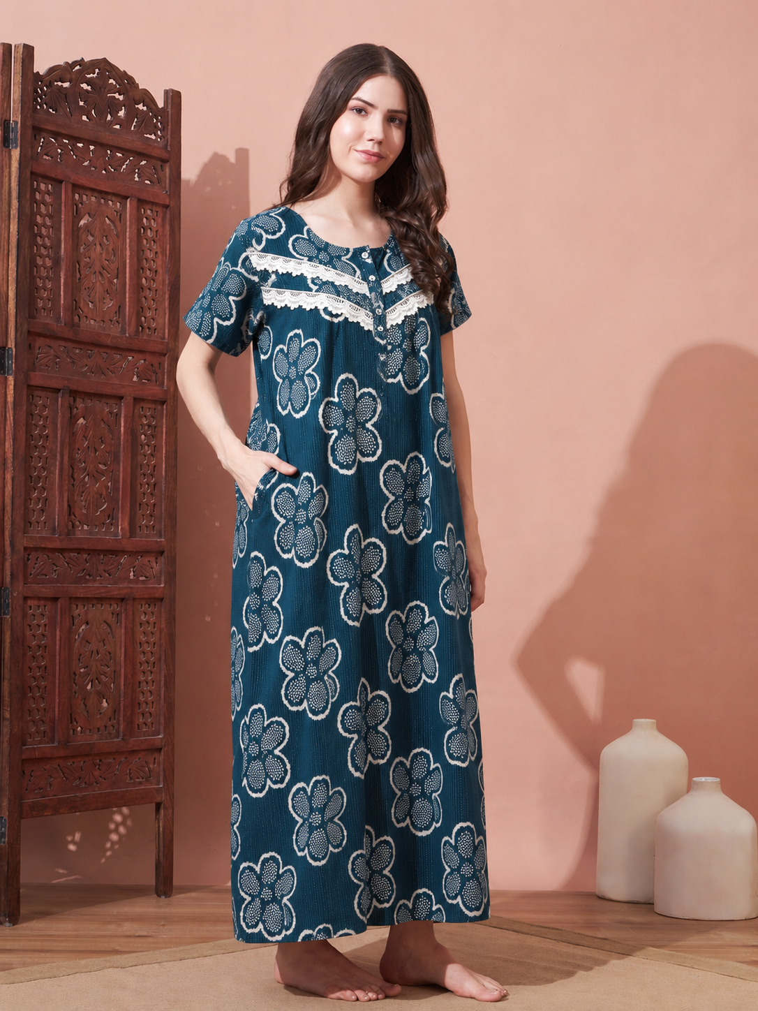A woman stands barefoot in a room wearing a long teal nightgown with a white floral block print pattern and lace trim at the neckline. She is posed next to a carved wooden screen, with two white ceramic vases on the floor against a peach-colored wall.