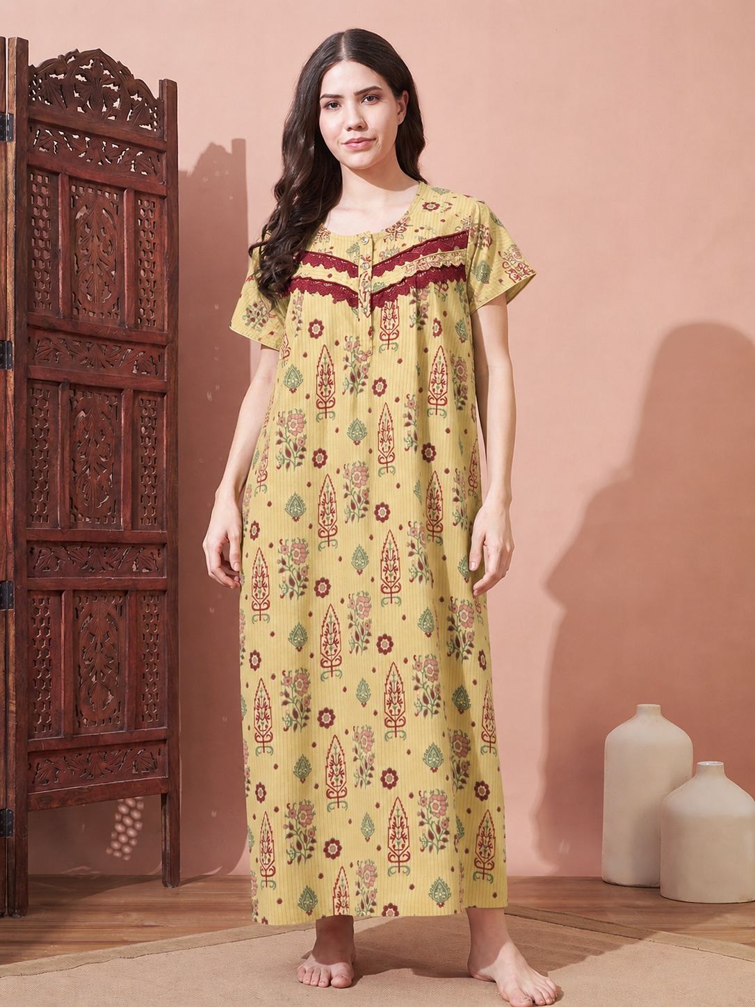 A woman stands barefoot in a bedroom-like setting wearing a long yellow printed nightgown with red and green motifs and a red embroidered neckline. A carved wooden screen is on the left, and two white vases sit on the floor against a peach wall.