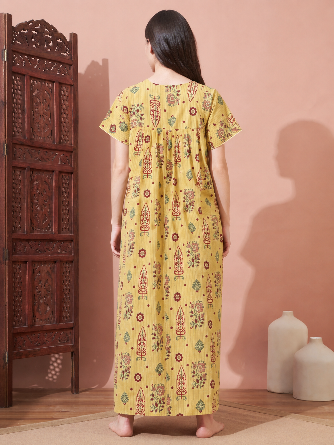 A woman stands with her back to the camera wearing a yellow short-sleeve maxi nightgown with a colorful hand-block print. The setting features a warm pinkish-beige wall, a dark carved wooden screen, and two white ceramic vases.