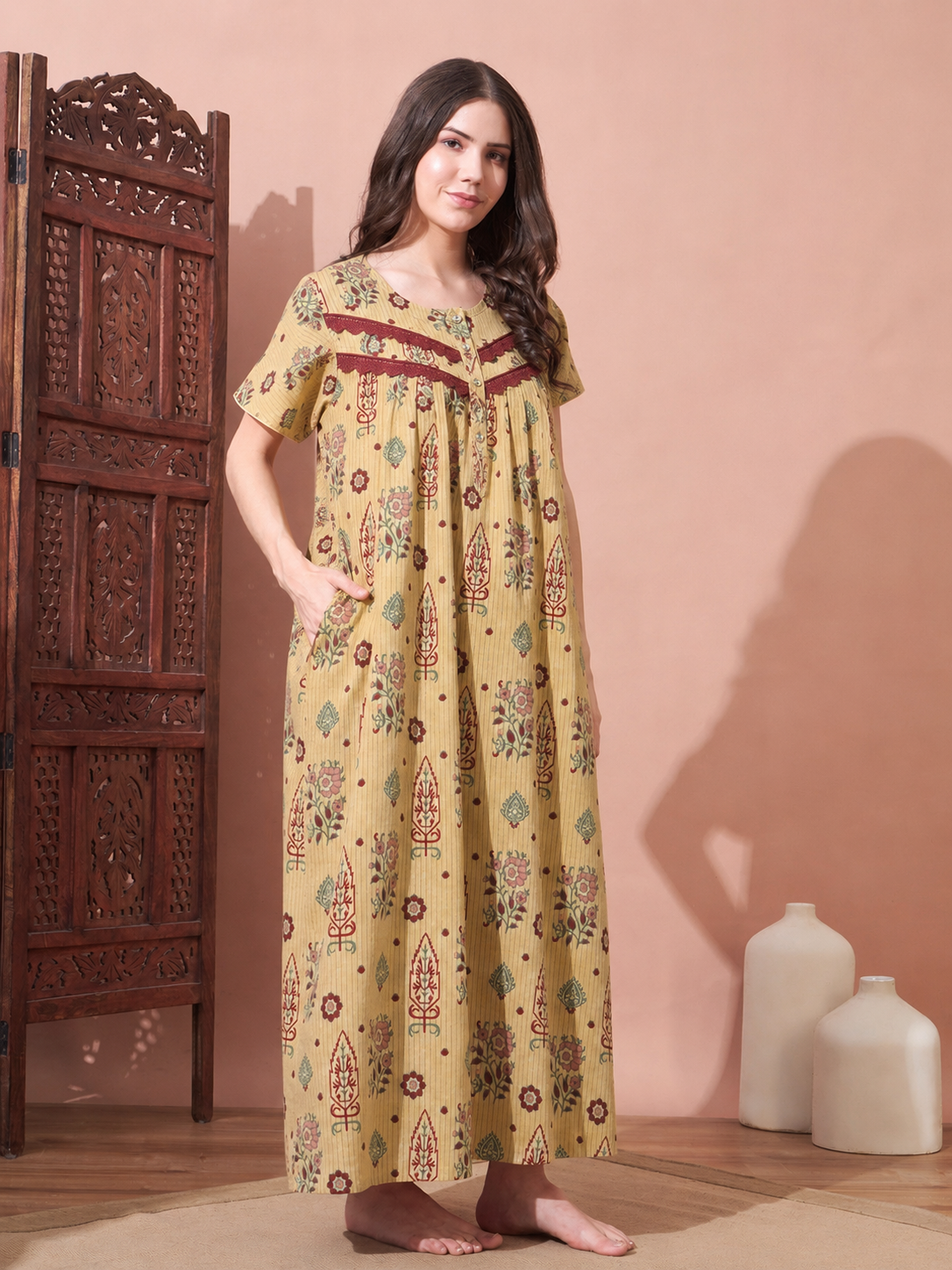 A woman stands barefoot in a room wearing a long, loose-fitting yellow printed nightgown with red and green motifs and a decorative neckline. She is posed next to a carved wooden screen, with two white ceramic vases on the floor against a warm pinkish-beige wall.