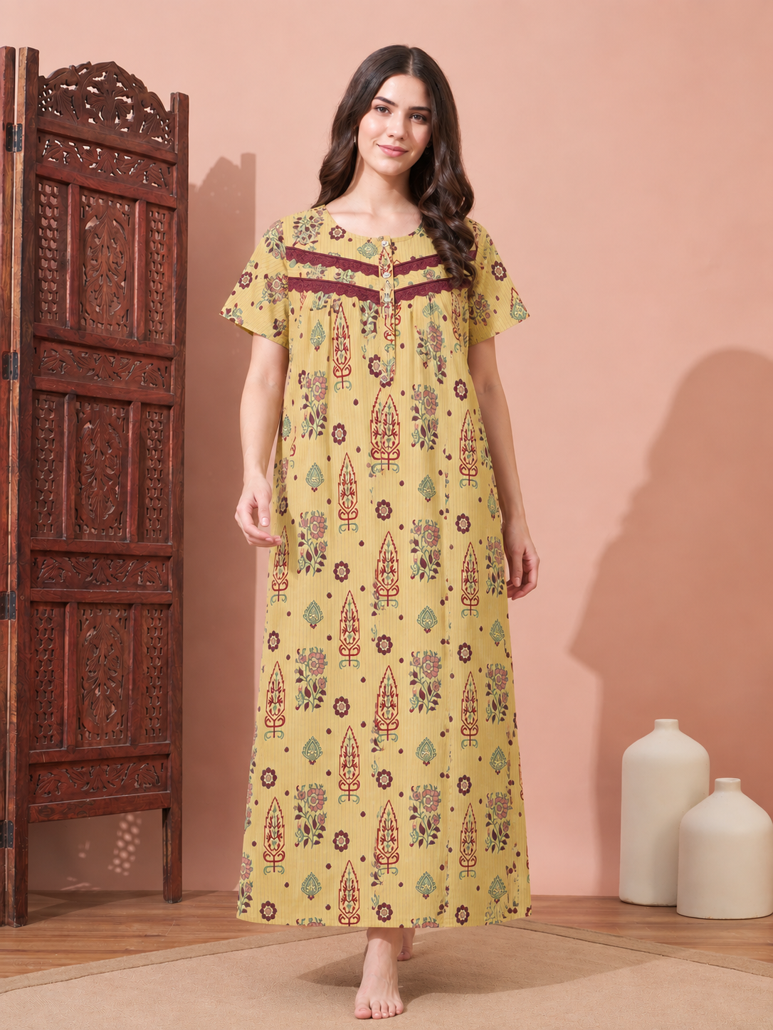 A woman stands barefoot indoors wearing a long yellow printed nightgown with short flutter sleeves and a decorative neckline. The dress has a multicolor floral and geometric pattern with brown and red accents.