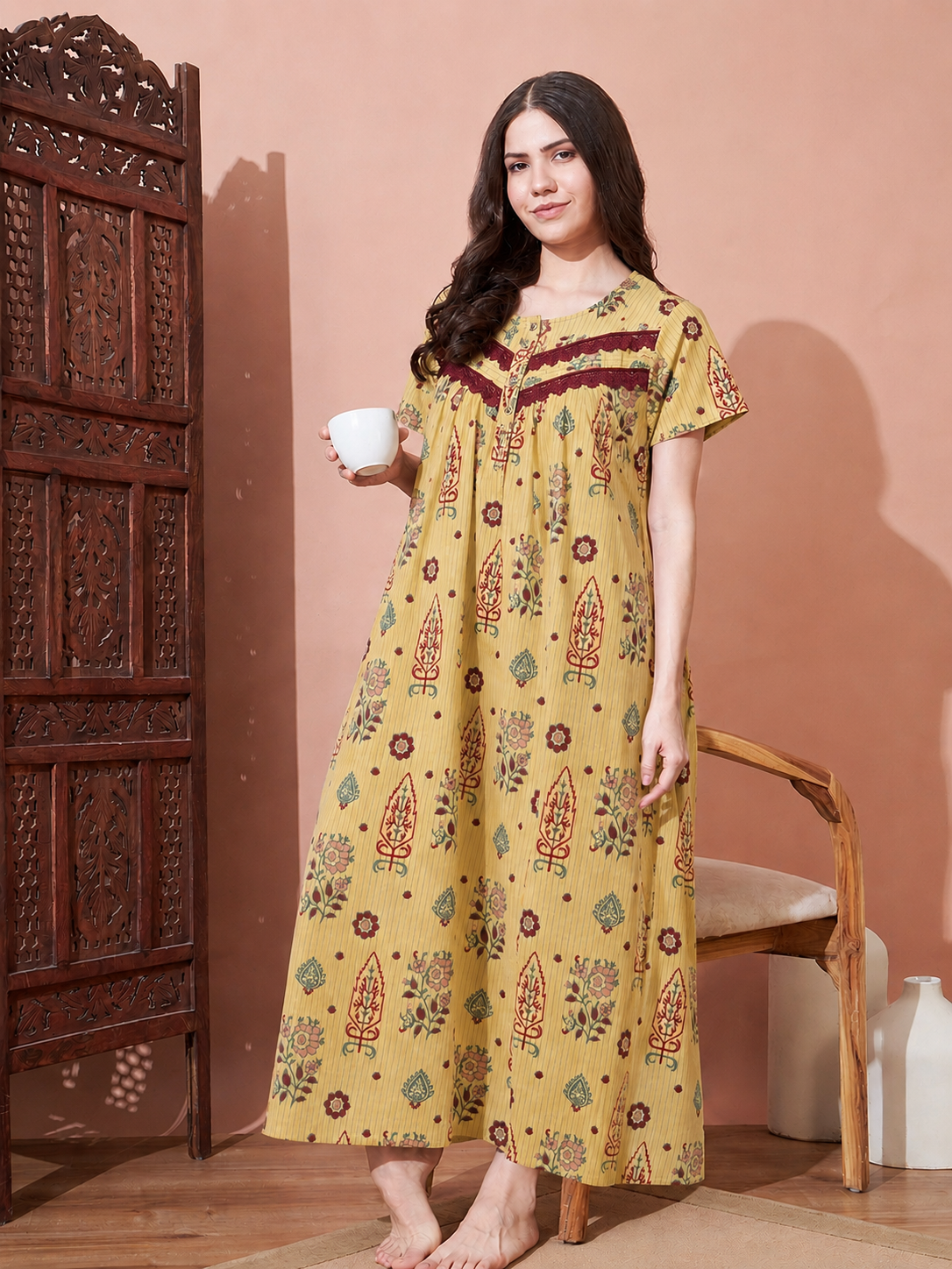 A woman stands barefoot indoors wearing a long yellow printed nightgown or kaftan dress with red and green motifs, holding a white cup. A carved wooden cabinet, chair, and peach wall are visible in the room.