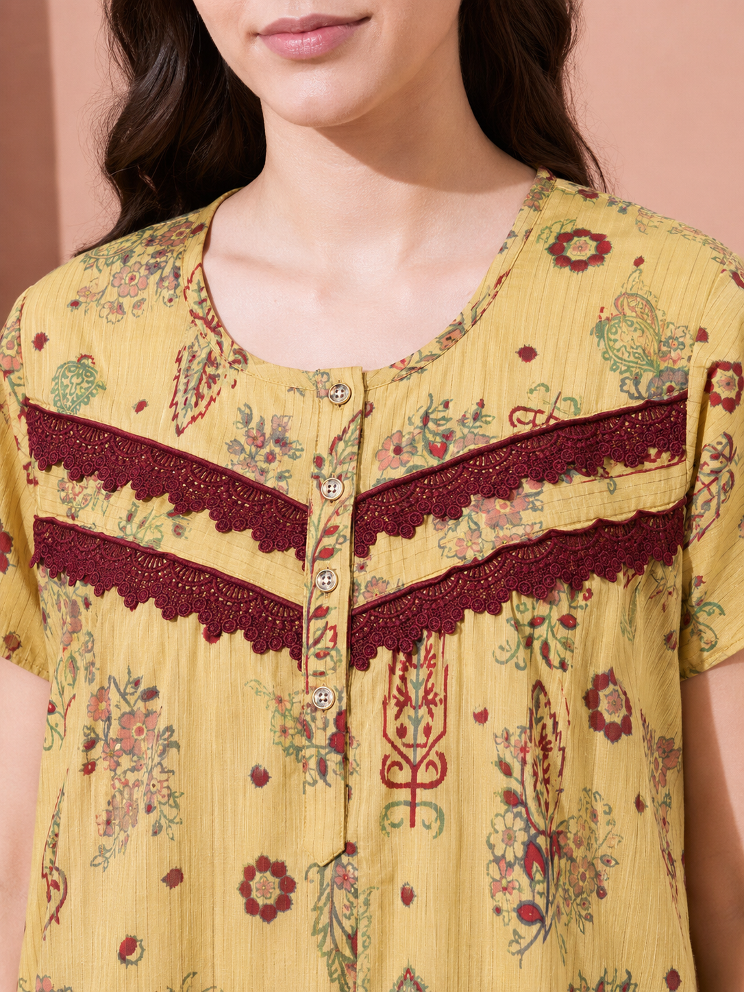 A close-up of a woman wearing a yellow short-sleeve top with a multicolor floral print and dark red lace trim across the chest.