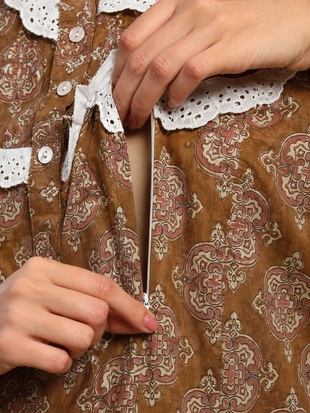 Close-up of a person's hands opening a zipper on a brown patterned dress with beige and pink damask motifs and white lace trim.