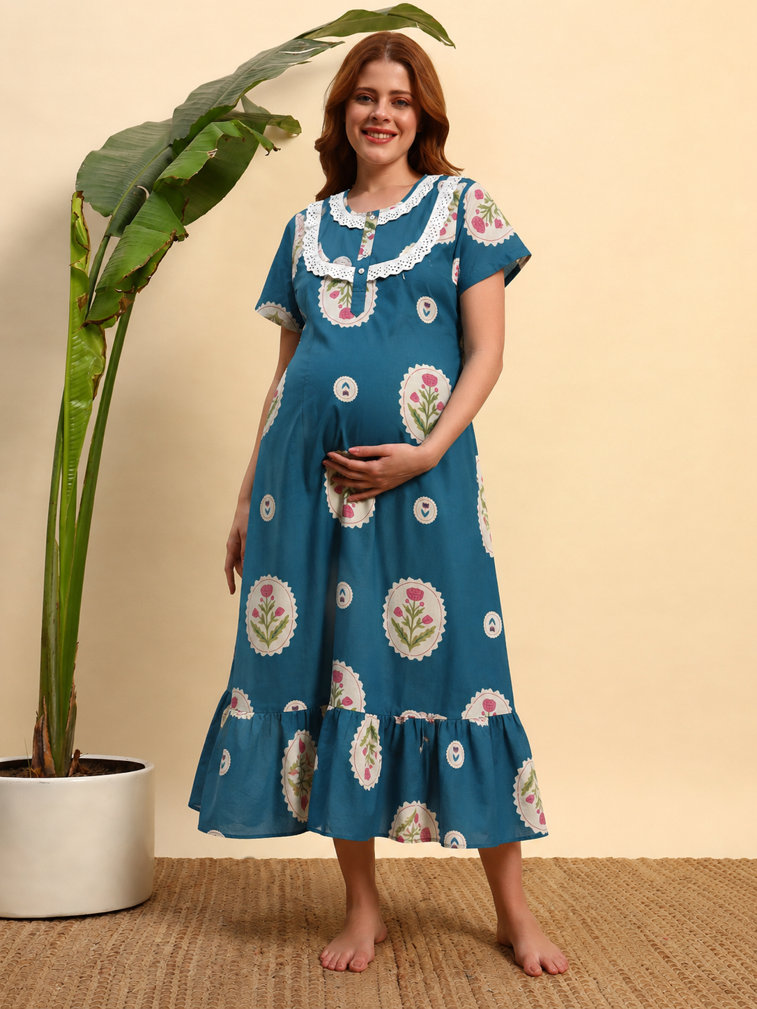 A pregnant woman wearing a teal blue maternity nightgown with white floral embroidery and ruffled trim, standing barefoot in a room with a beige wall and a large potted plant.