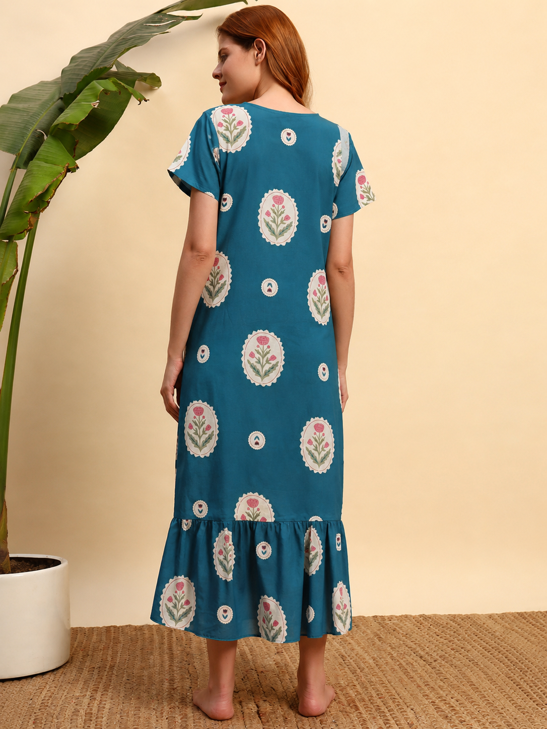 A woman stands barefoot in a studio setting wearing a teal blue midi dress with a ruffled hem and short sleeves. The dress features a white and pink floral block print pattern.