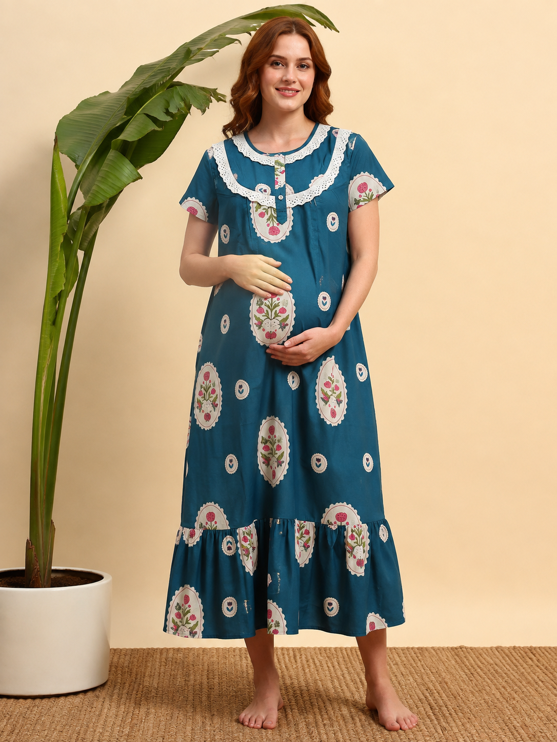 A woman wearing a teal blue maternity nightgown with white floral embroidery and white lace trim, standing barefoot in front of a beige wall next to a potted plant.