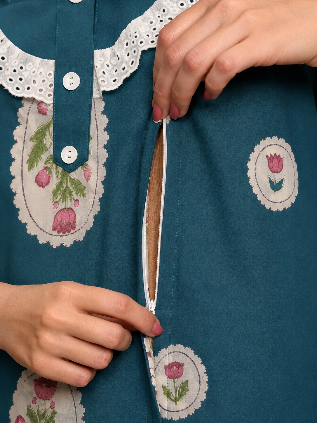 Close-up of a teal blue dress with floral embroidered patches and white eyelet lace trim, showing a person's hands opening a front zipper.