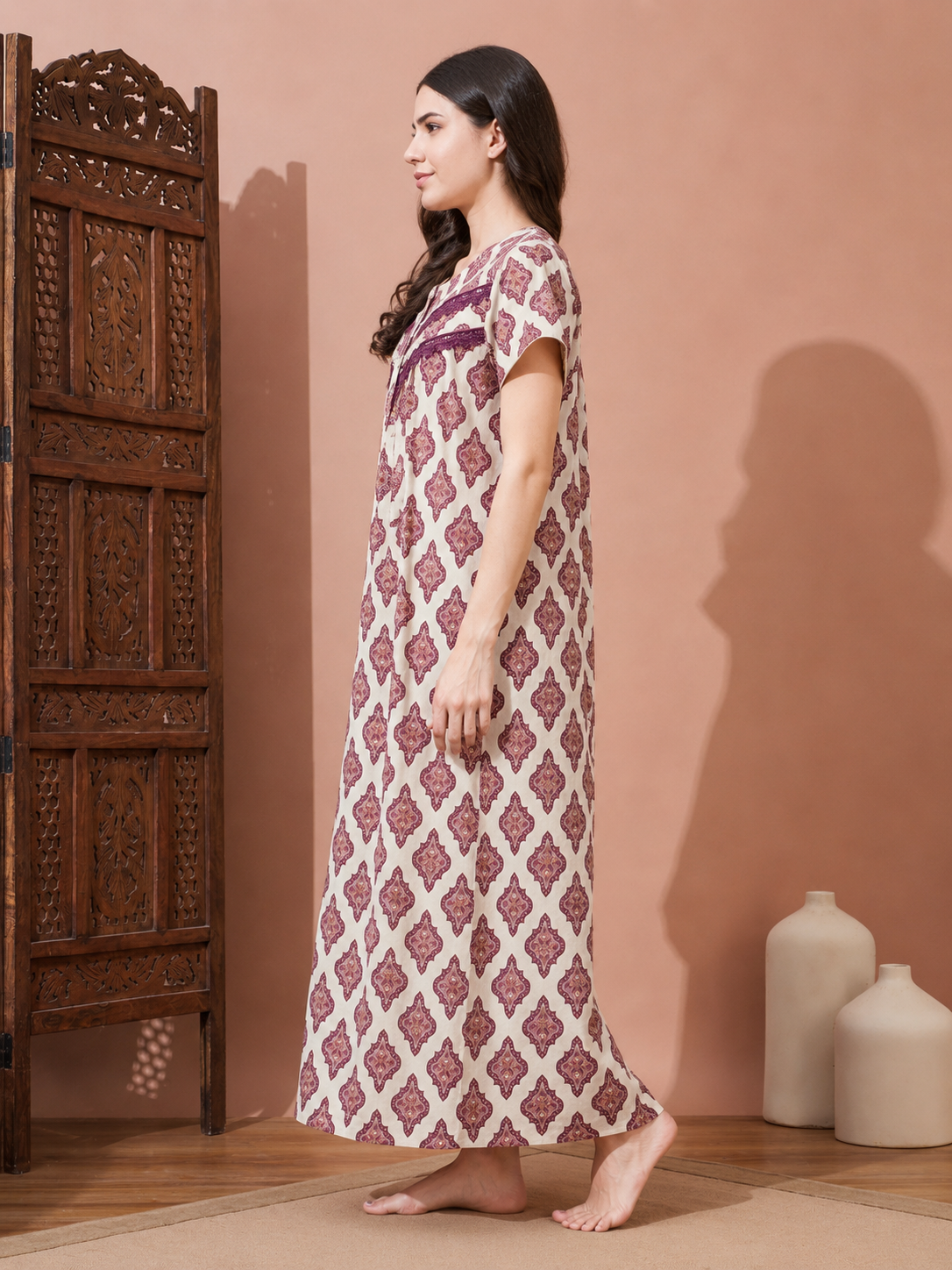 A woman stands barefoot in profile wearing a long, short-sleeve nightgown with a pink and cream geometric pattern, set against a warm peach-colored wall with a carved wooden screen and decorative vases.