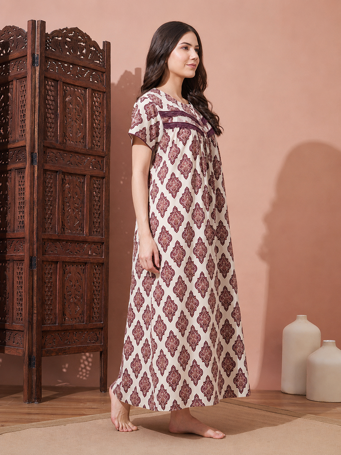 A woman stands barefoot in a studio setting wearing a long cream nightgown with a dusty rose geometric diamond pattern and matching embroidered chest detail. A carved dark wood screen is on the left, and two white ceramic vases sit on the floor against a warm peach-pink wall.