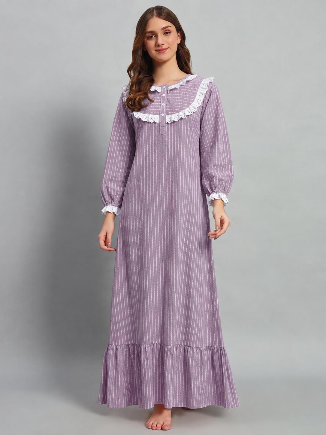 A woman modeling a long-sleeved lavender and white vertically striped nightgown with white ruffle trim on the collar and cuffs, standing against a solid gray studio background.