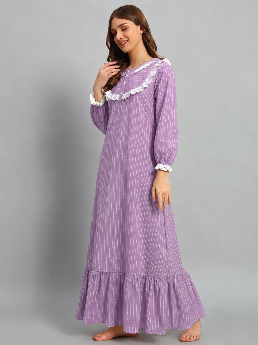 A woman modeling a long-sleeved lavender nightgown with white ruffled trim at the collar and cuffs, featuring a subtle vertical stripe pattern and a ruffled hem, set against a solid gray studio background.