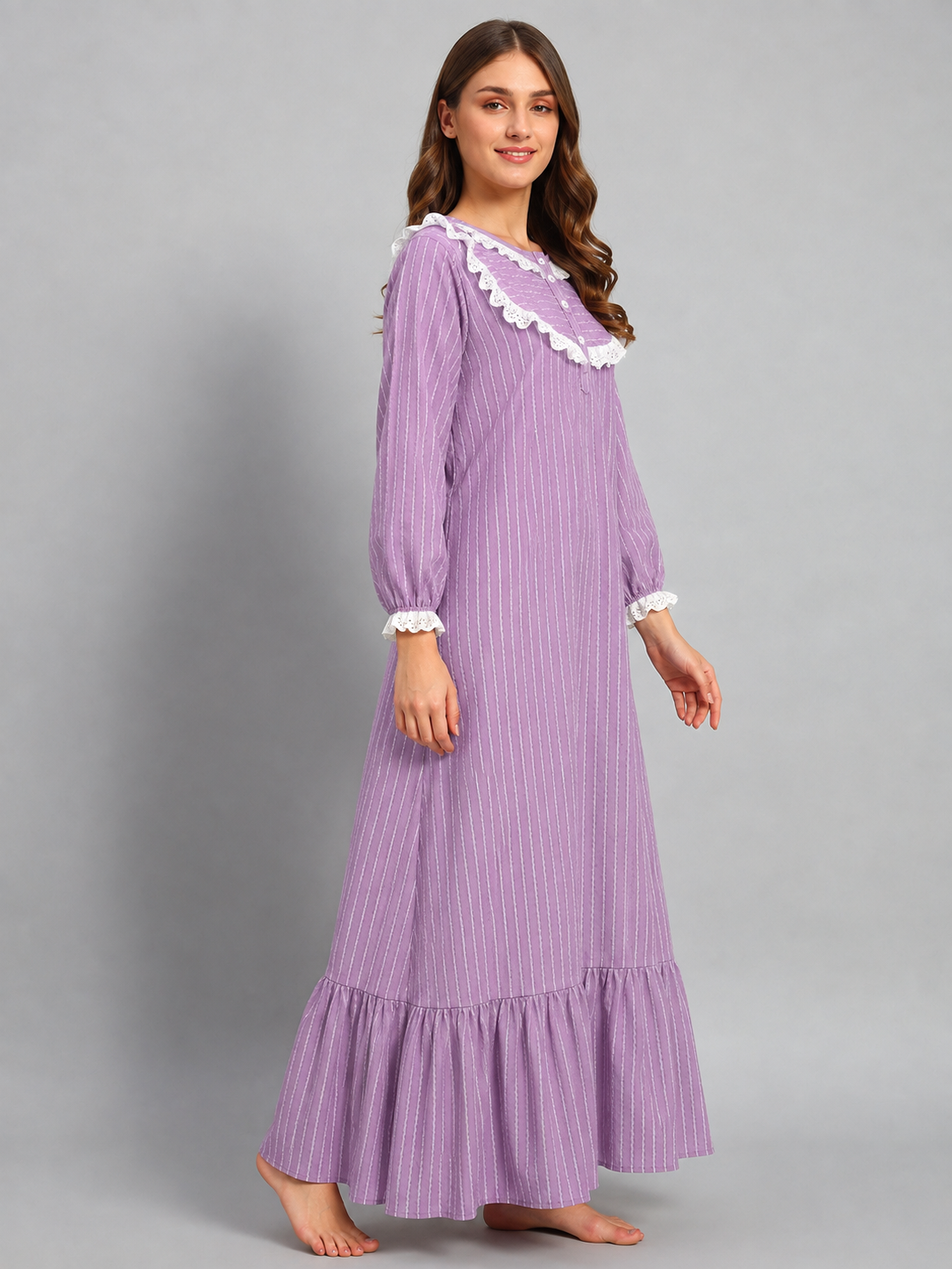 A woman modeling a long-sleeved lavender nightgown with white ruffled trim at the collar and cuffs, set against a solid gray studio background.