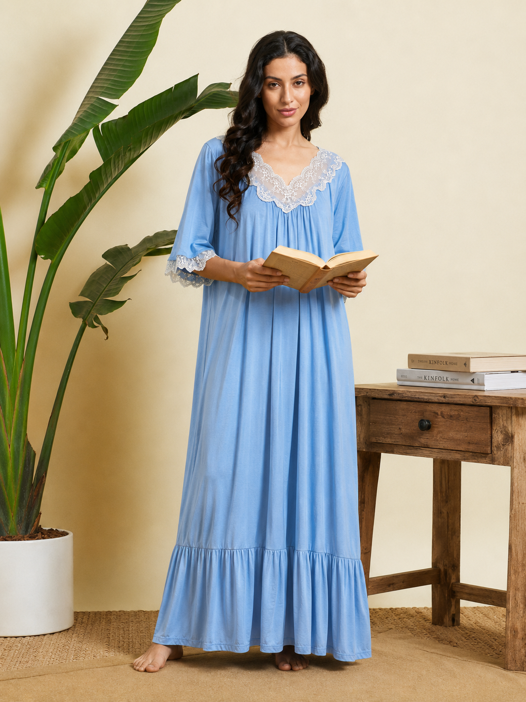 A woman stands barefoot in a bedroom-like setting wearing a long light blue nightgown with white lace trim at the neckline and cuffs, holding an open book. A potted plant, beige wall, and wooden side table with books are visible.