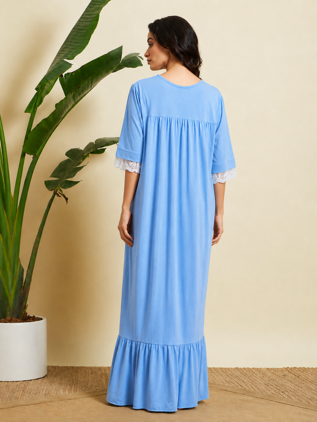 A woman wearing a light blue maxi dress with white lace trim on the sleeves, shown from the back in a studio setting with a potted plant and beige wall background.