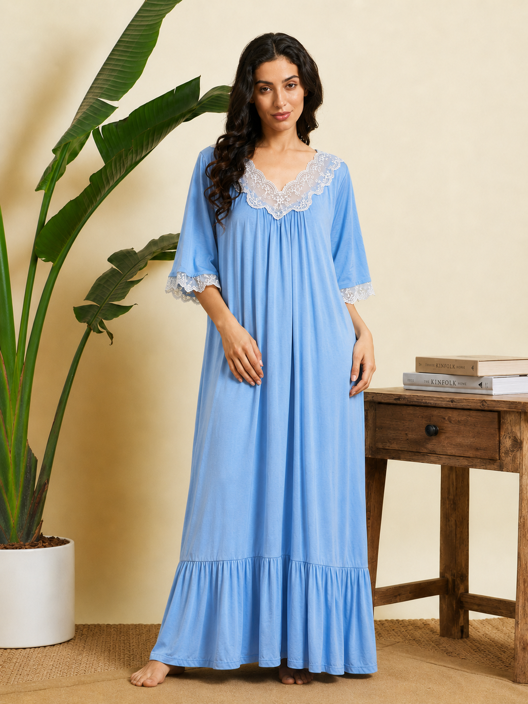 A woman stands barefoot in a bedroom-like setting wearing a light blue maxi nightgown with white lace trim at the neckline and sleeve cuffs. A potted plant and a wooden table with books are visible in the background.