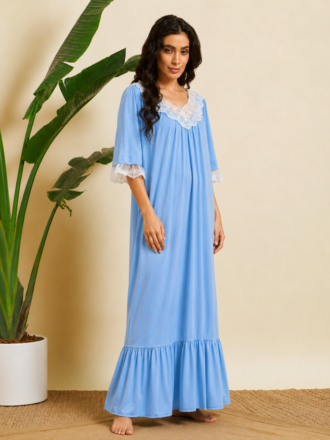 A woman stands barefoot indoors wearing a light blue maxi nightgown with white lace trim at the neckline and sleeve cuffs. She is posed next to a large potted plant against a warm beige wall.