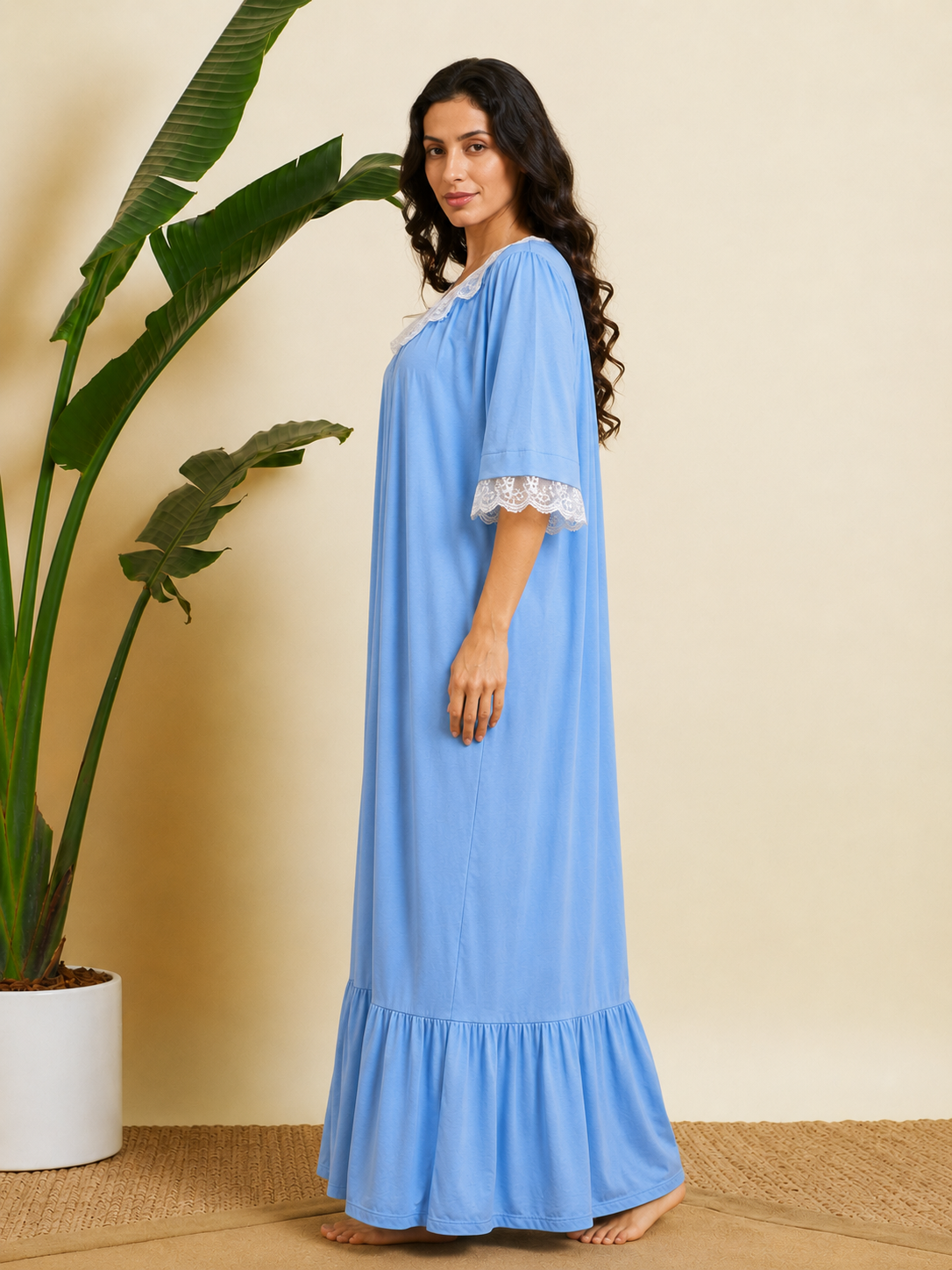 A woman wearing a light blue maxi nightgown with white lace trim on the collar and sleeve cuffs, standing barefoot in a room with a beige wall, a potted plant, and a woven rug.