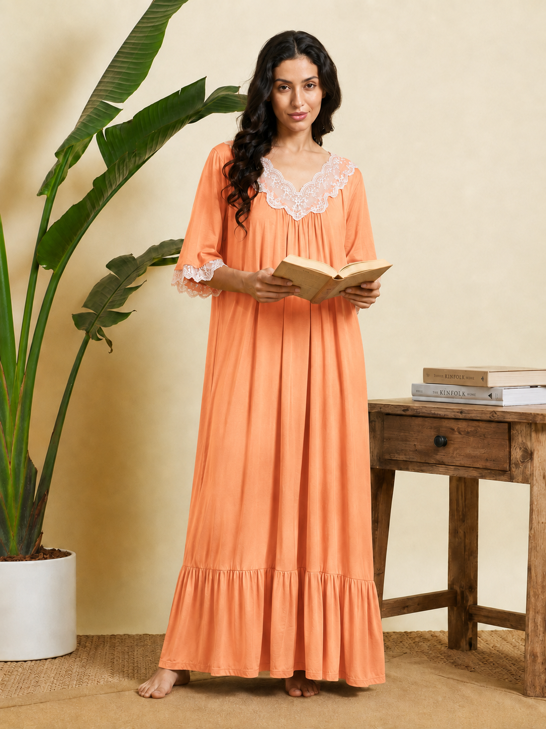 A woman stands barefoot in a warm-toned room wearing a long peach maxi nightgown with white lace trim at the neckline and cuffs, holding an open book. A potted plant and a wooden side table with books are visible in the background.